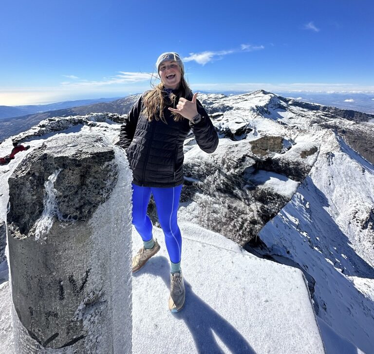 mackenzie stands on top of snowy mountain with blue sky behind her